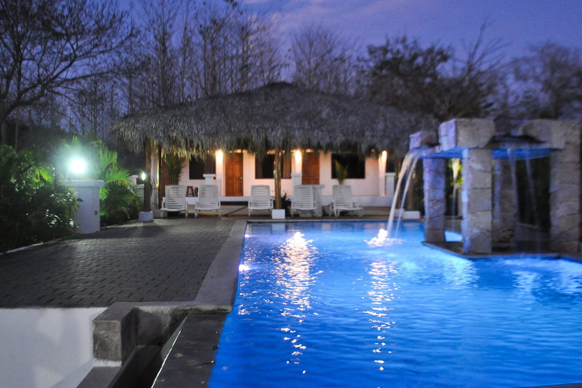 A swimming pool with a waterfall feature, surrounded by lounge chairs and a thatched-roof structure.