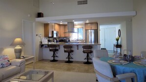 A modern kitchen with a central island, bar stools, and a dining area with a table set for two. - Palm Desert retreat right on the golf course with King en-suite  (Palm Desert)