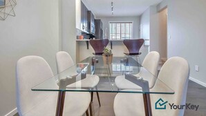 A modern dining area with a glass table and white chairs, a kitchen in the background, and a window with blinds. - Enchanted Escape in Northwest Calgary Townhome (Calgary)