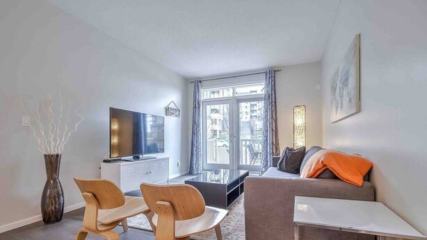 A modern living room with a sofa, two wooden chairs, a TV, and a large window with a view of buildings. - Enchanted Escape in Northwest Calgary Townhome (Calgary)