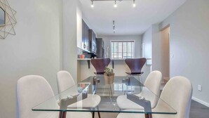 A modern dining area with a glass table and white chairs, a kitchen in the background, and a window with blinds.