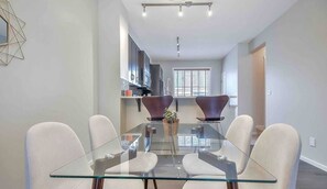 A modern dining area with a glass table and white chairs, a kitchen in the background, and a window with blinds. - Enchanted Escape in Northwest Calgary Townhome (Calgary)