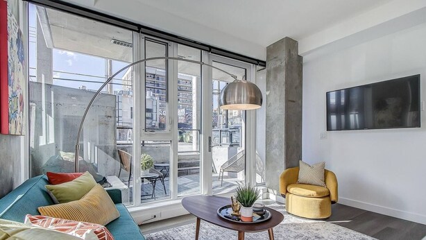 A modern living room with a large window, a sofa, a yellow armchair, and a TV mounted on the wall.