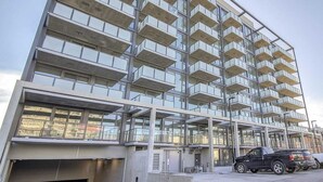 A modern multi-story building with a glass facade, multiple balconies, and a parking area with vehicles.