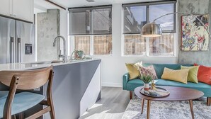 A modern kitchen with a large island, a dining area with a wooden chair and table, and a living room with a teal sofa and colorful cushions.