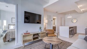 A modern living room with a flat-screen TV, a sofa, a coffee table, and a dining area with bar stools.