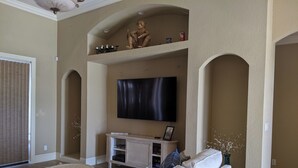 A living room with a large flat-screen TV mounted on the wall, a white sofa with blue cushions, a small round coffee table, and a white console table with decorative items. - Cape Coral Paradise: Executive Home, weekly & monthly rates upon request. (Cape Coral)