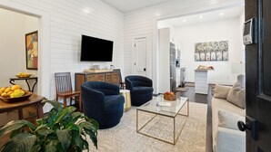 A modern living room with a flat-screen TV, two blue armchairs, a glass coffee table, and a beige sofa. - 8B at Saint Philip Square, One Block to King, Spacious Group and Family Favorite (Charleston)