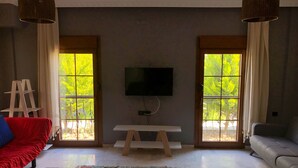 A living room with a TV, a bench, and a sofa. There are two windows with curtains, and a colorful rug on the floor.