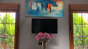 A room with a TV mounted on the wall, a vase with flowers on a table, and a view of greenery through the windows.