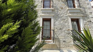A stone building with three windows and balconies, surrounded by greenery.