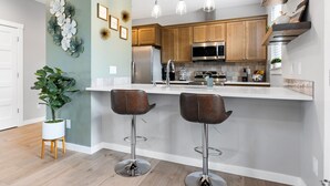 Une cuisine moderne avec des meubles en bois, des appareils en acier inoxydable et un plan de travail blanc. On y trouve deux tabourets de bar et une plante en pot.