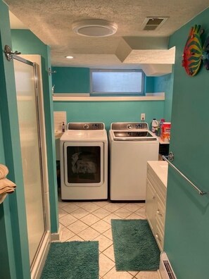 Compact laundry room with two front-loading washing machines and a sink, featuring a shower door and ceiling vent.