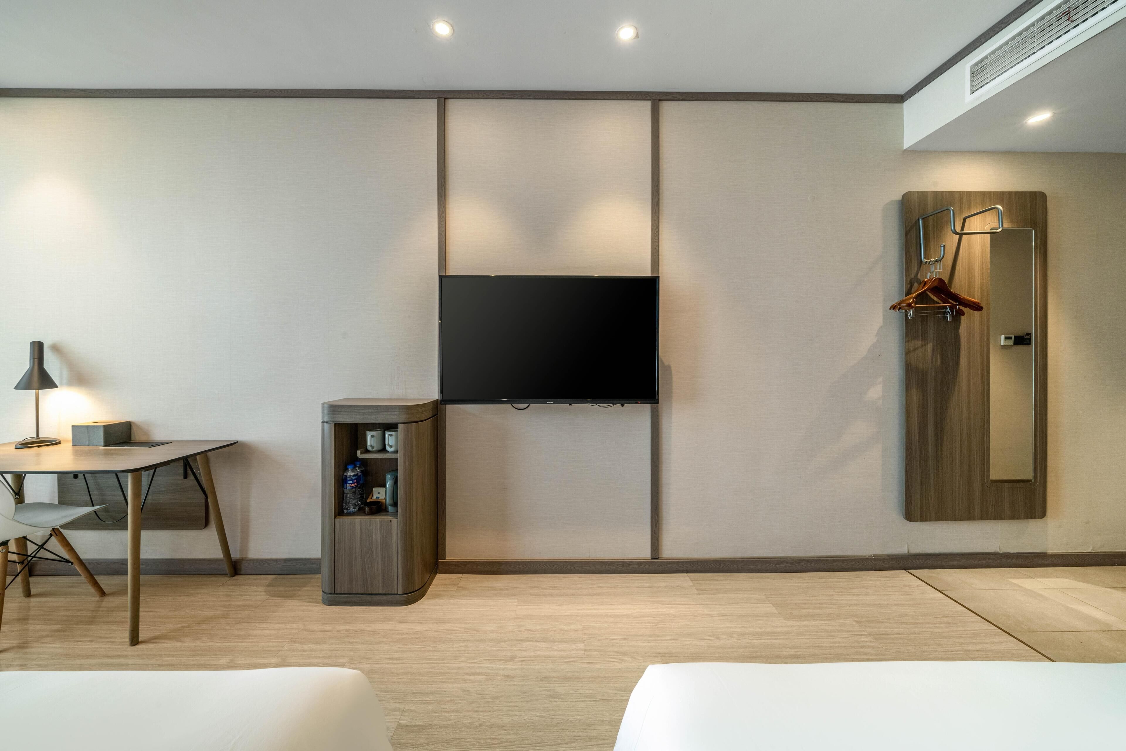 Una habitación de hotel moderna con un televisor de pantalla plana en la pared, una mesita con lámpara, un minibar y un perchero.
