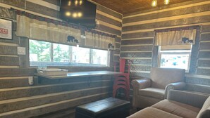 Una habitación acogedora de estilo cabaña con un ventanal grande, un televisor de pantalla plana montado en la pared y una zona de estar confortable con sofá y reposapiés.