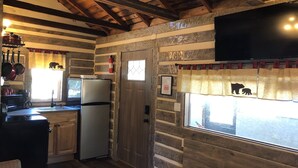 Interior rústico de una cabaña con cocina, paredes de madera, televisor montado y una ventana con cortinas que muestran siluetas de osos.