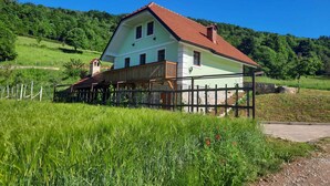 Una casa de dues plantes amb teulada de rajola vermella, envoltada de camps verdosos i una tanca de fusta.