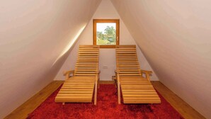 A small attic room with two wooden chairs, a red carpet, and a window with a view of greenery.