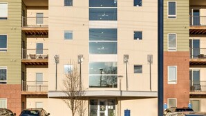 Un immeuble à plusieurs étages doté d’une entrée centrale, d’un espace de stationnement et surplombant un ciel dégagé.