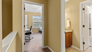 A hallway with beige walls and white trim, leading to a room with a desk and office chair, and another room with a sink and countertop.