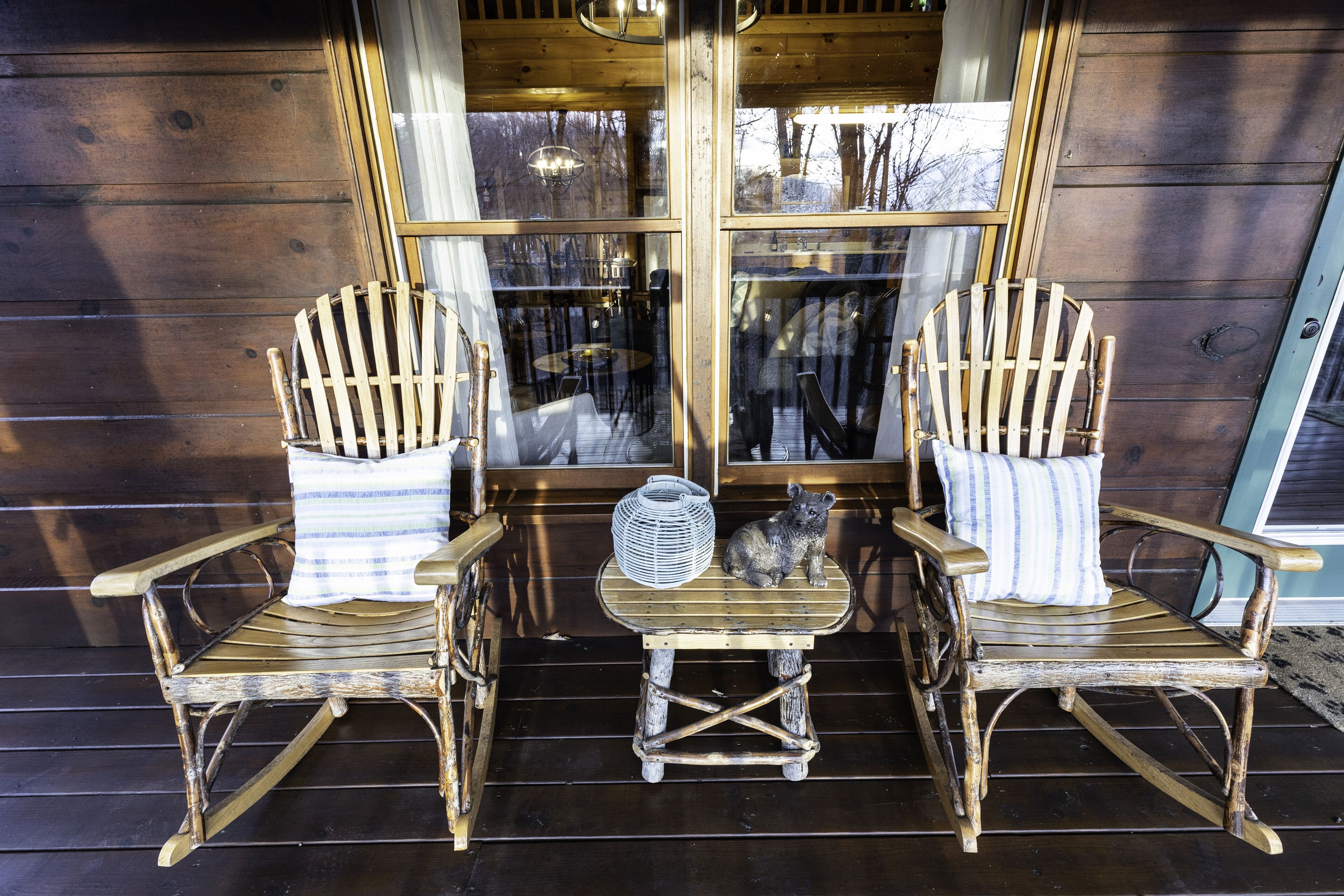Two rocking chairs with a small table and a fan between them, positioned on a wooden deck with a view of the interior through large windows.