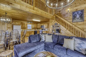 A wooden cabin interior with a sofa, dining table, chairs, kitchen area, and a staircase. - MTN Cabin Sleeps 10 - Camping & Fishing - Family Vacay & Outdoor Adventures (Ellijay)