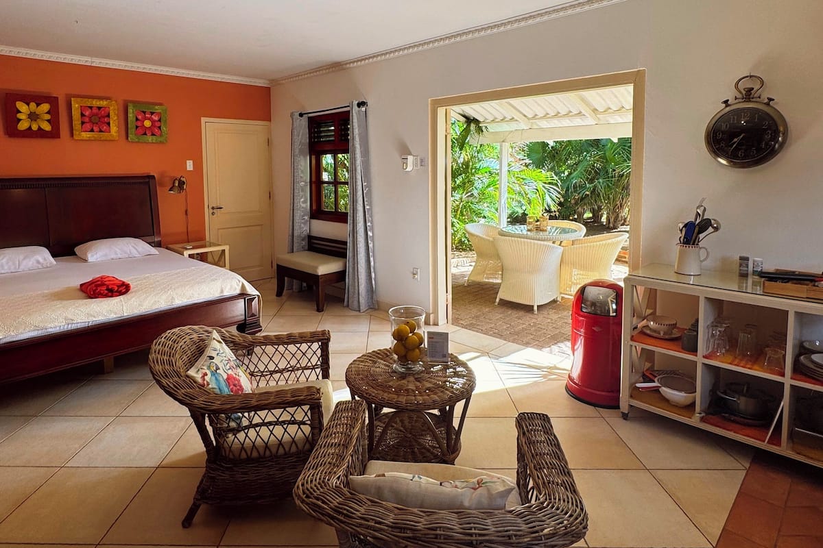 A bedroom with a bed, two wicker chairs, a small table with a vase, a red trash bin, and a shelf with kitchenware.
