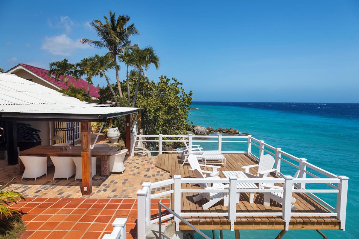 A wooden deck with white chairs and a table, overlooking turquoise ocean waters.