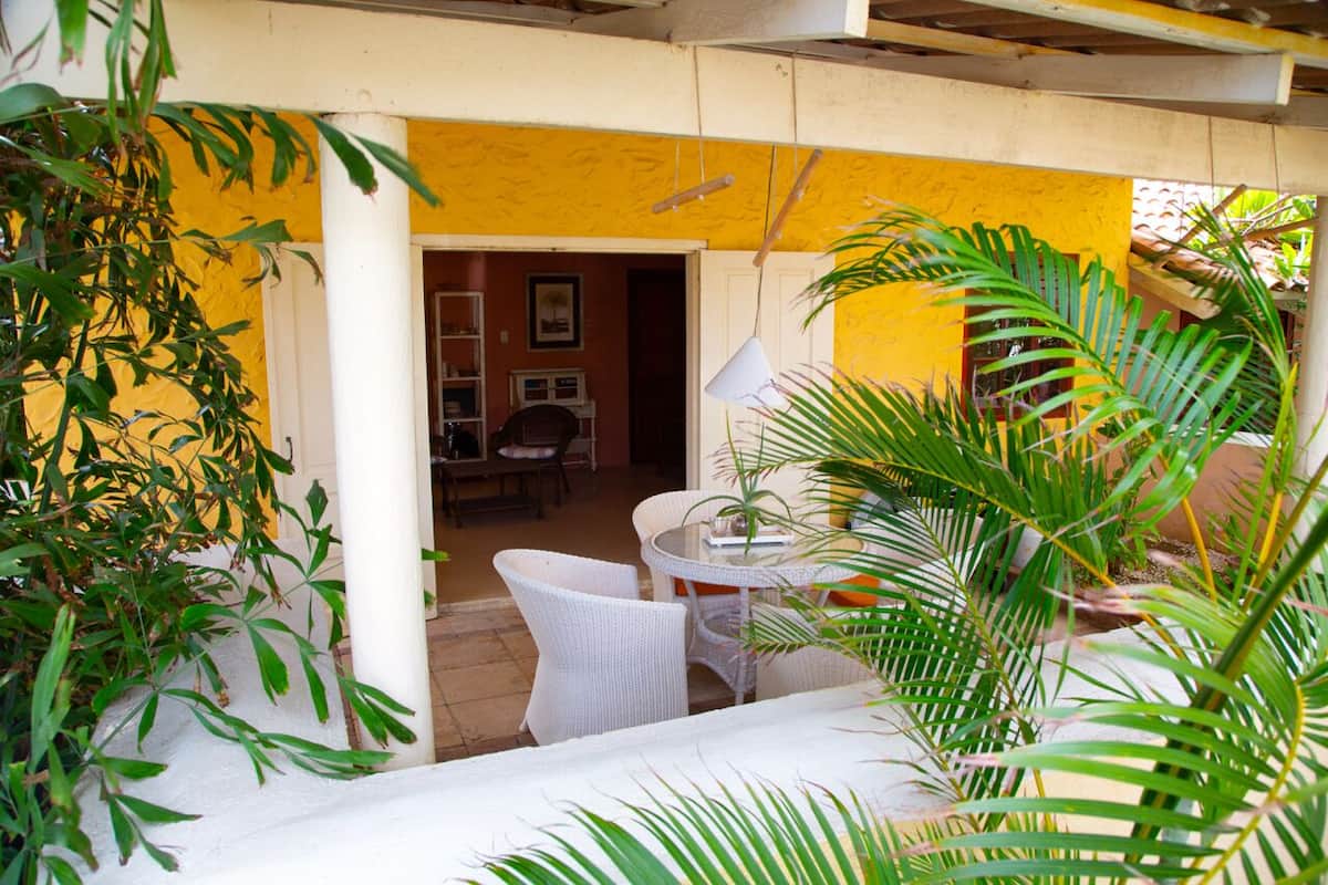 A patio area with a white wicker table and chairs, surrounded by potted plants and a yellow wall.