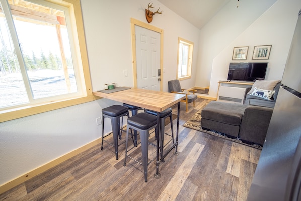 A modern interior with a wooden dining table, bar stools, a gray sofa, and a television.