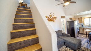 A staircase with wooden steps and a white wall. There is a ceiling fan and a kitchen area visible in the background.
