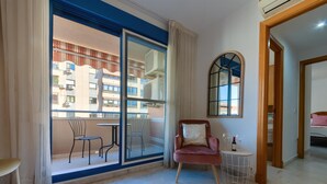 Una sala moderna con un amplio ventanal corredera que da a un balcón, un sillón rosa, una pequeña mesa redonda y una cama en la habitación contigua.