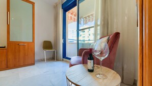 Una habitación con un armario de madera, una silla, una mesa con una botella de vino y una copa, y vistas a edificios a través de una ventana.