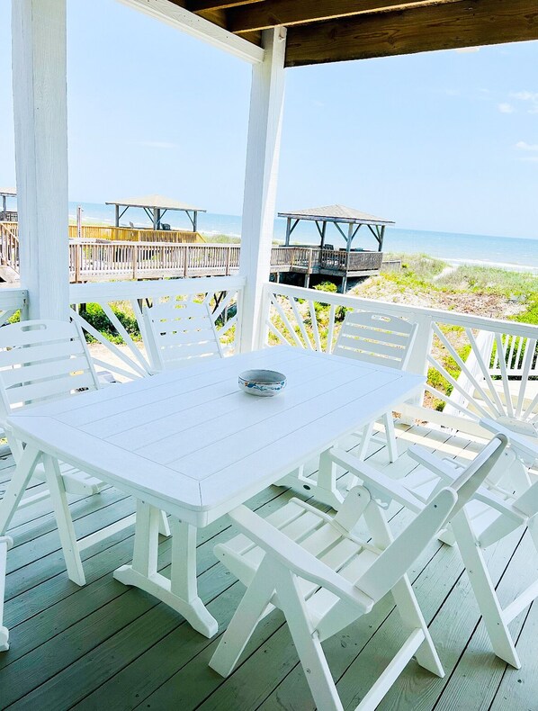 A white wooden deck with a table and chairs, overlooking a beach with wooden structures and a clear sky.