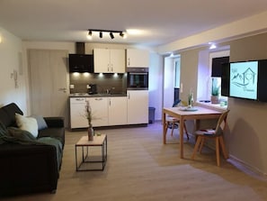 A modern living room with a sofa, a coffee table, and a dining area with a table and chairs. A kitchen with white cabinets and appliances is visible in the background.