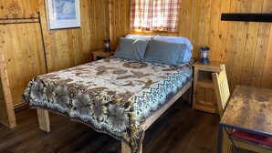 A wooden cabin bedroom with a bed, nightstands, a table, and a window with curtains. - THE FRAN - WALK EVERYWHERE IN DOWNTOWN CREEDE! 1 bedroom, 1 bath (SLEEPS 2) (Creede)