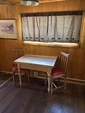 A wooden dining table with a red cushioned chair, a window with a patterned curtain, and a framed picture on the wall.