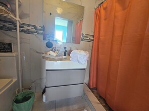 Bathroom features a sink, mirror, and shower, with toiletries and a trash can nearby.