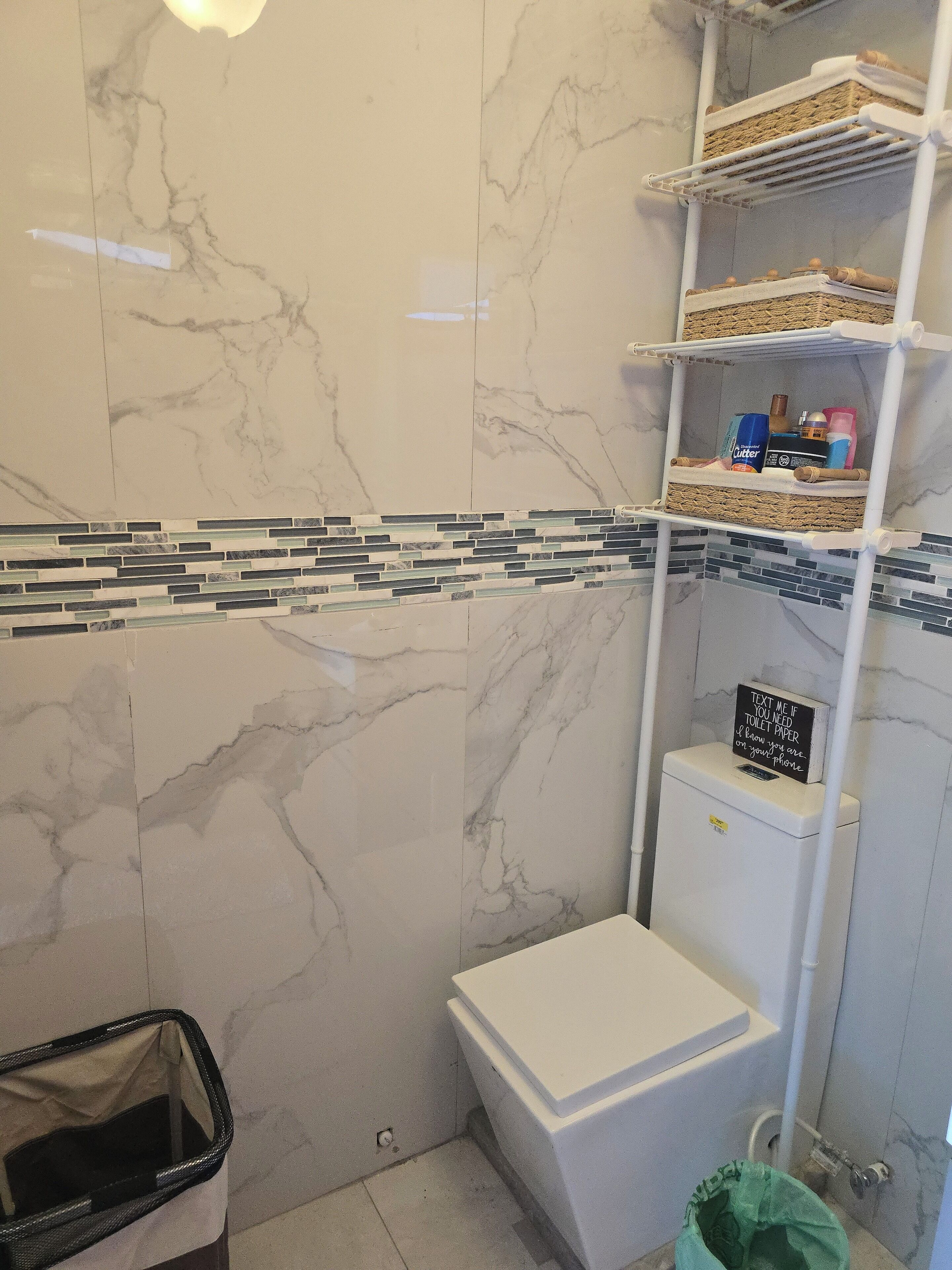 A bathroom with a marble wall, a white toilet, a shelf with toiletries, and a trash bin.