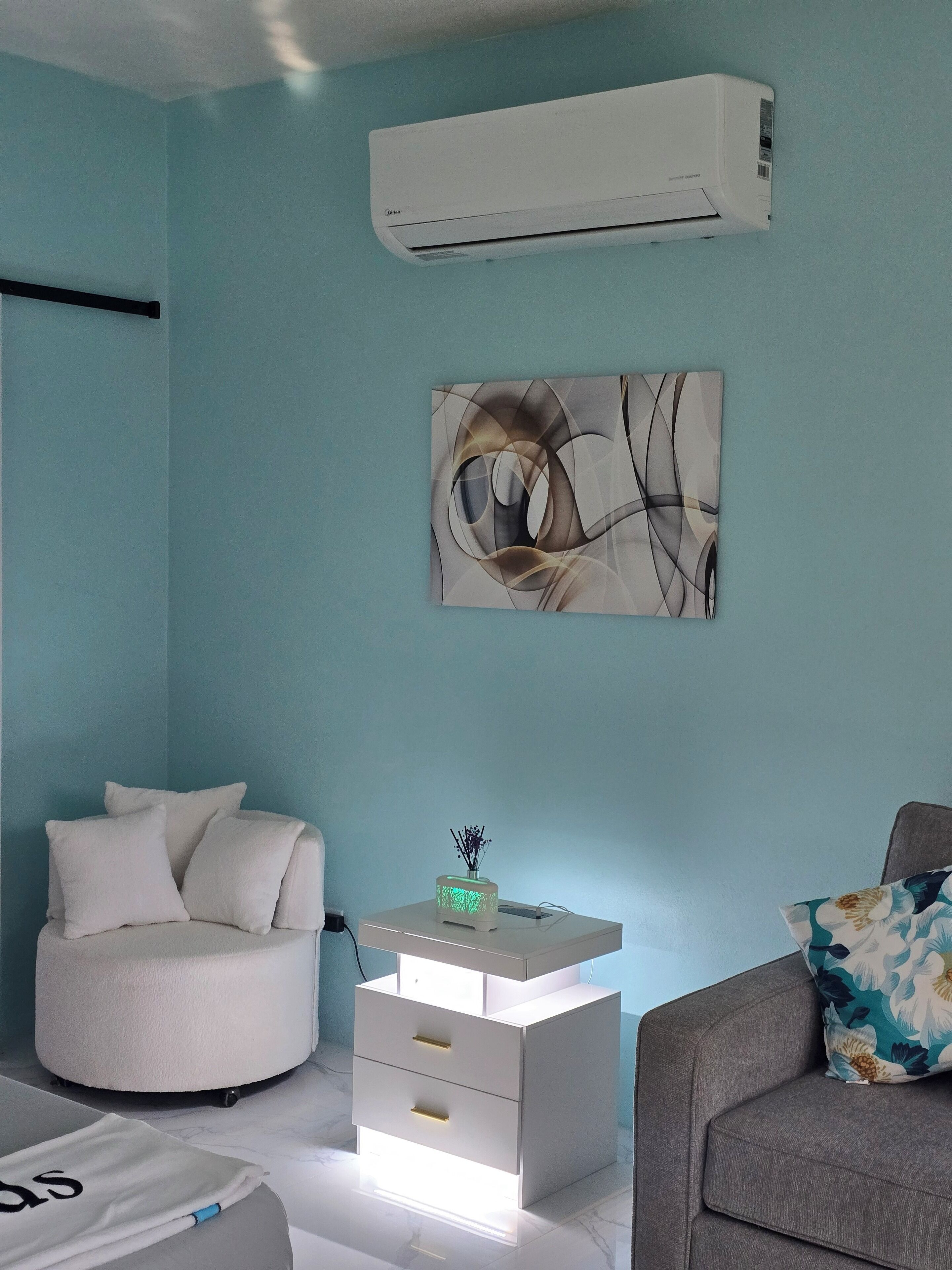A modern living room with a teal wall, a white air conditioner, a round white chair, a white bedside table with drawers, and a gray sofa with a floral pillow.