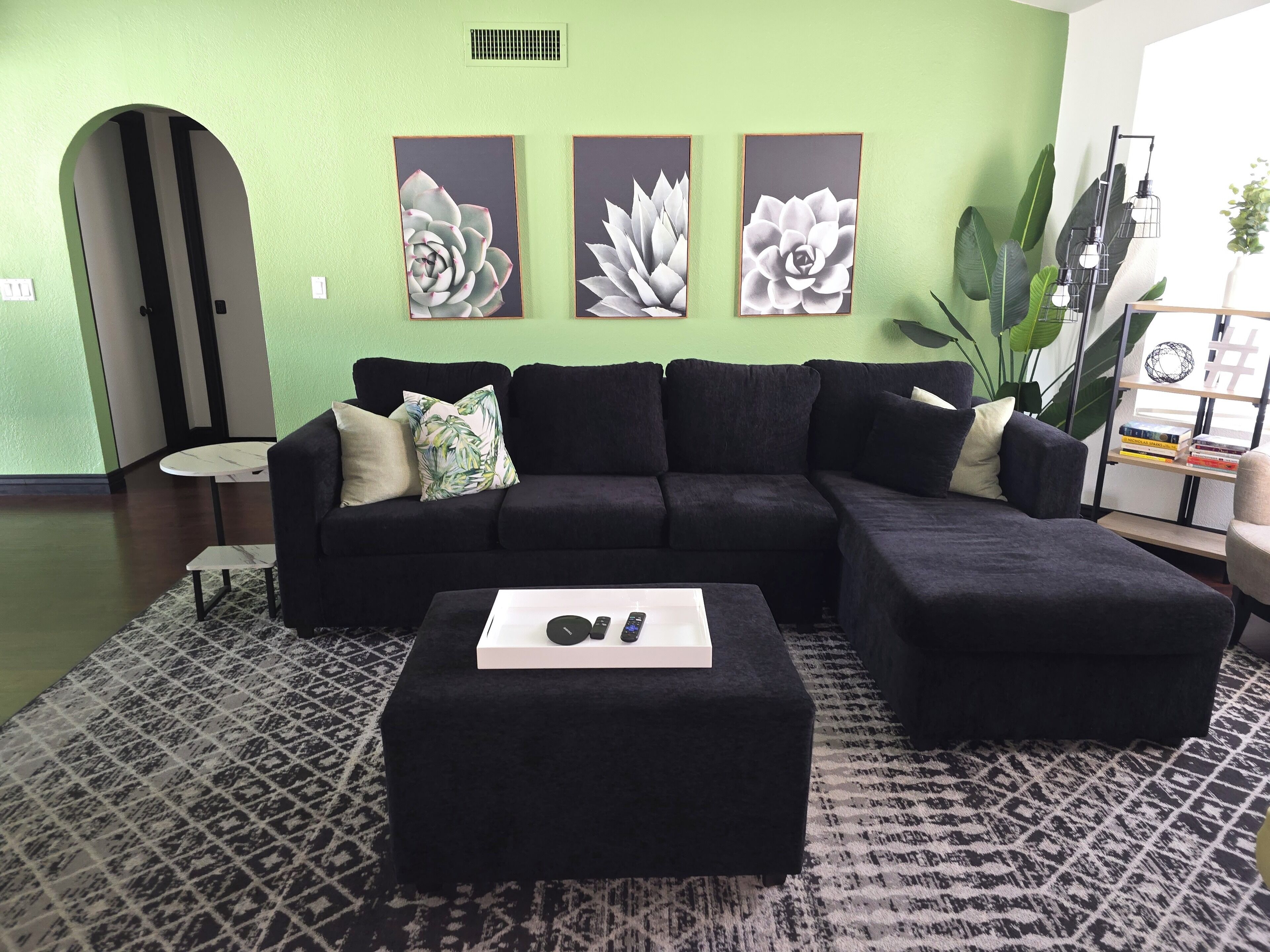 A living room with a black sectional sofa, a small round table, and a black ottoman with a remote control. There are three framed floral paintings on the wall and a large green plant in the corner.