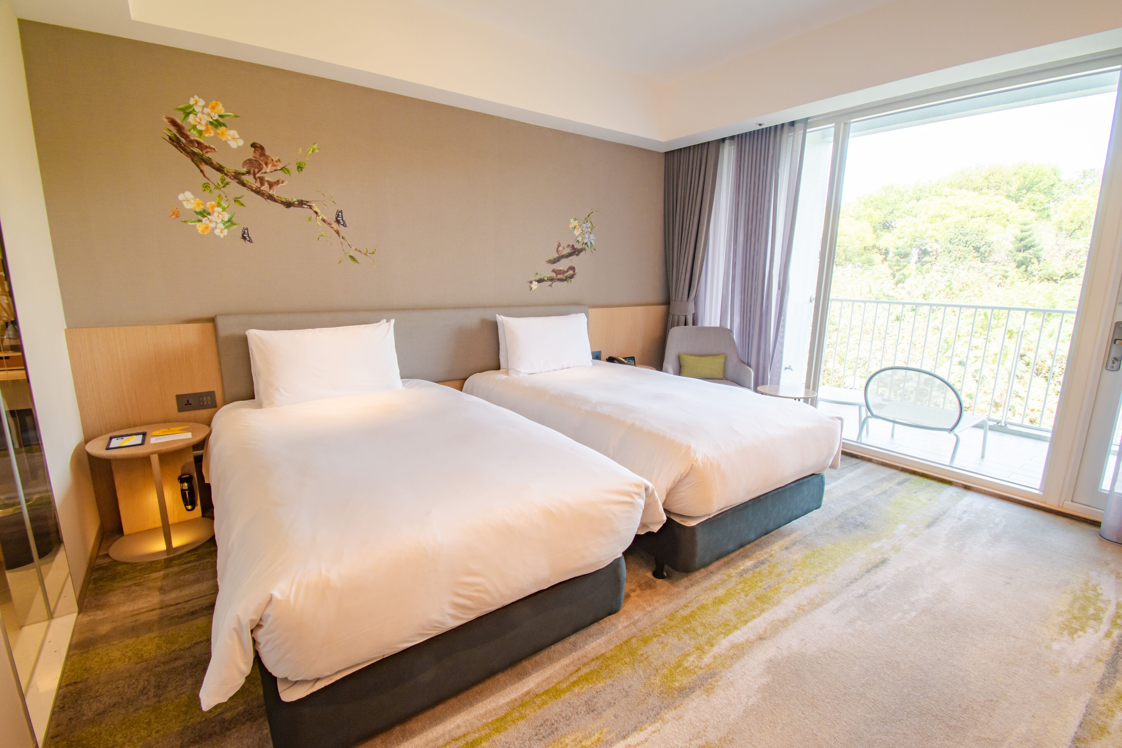 Habitación de hotel con dos camas, una mesita, una silla y un balcón con vistas a zonas verdes.