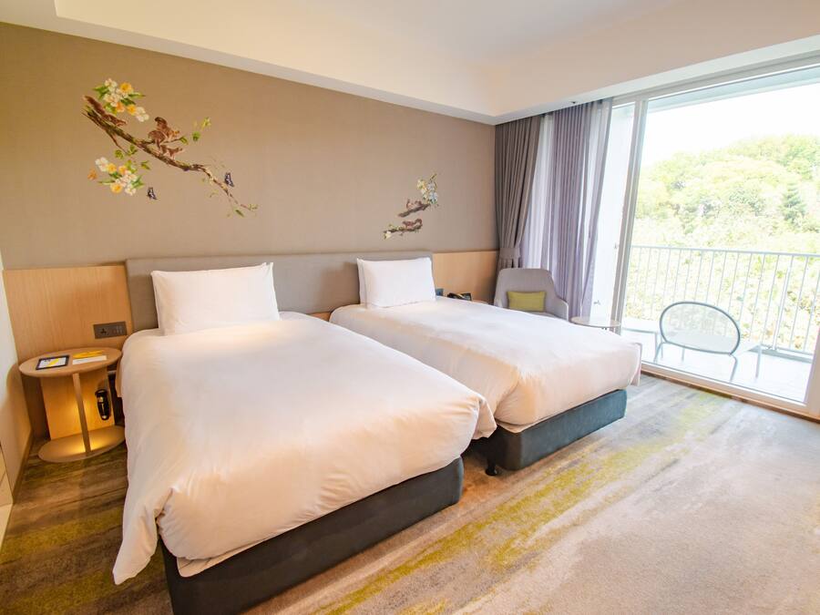 A hotel room with two beds, a small table, a chair, and a balcony with a view of greenery.