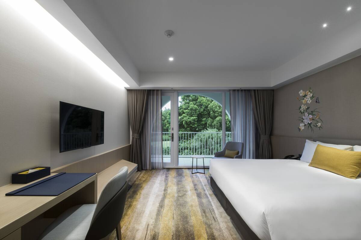 Ein modernes Hotelzimmer mit einem großen Bett, einem Schreibtisch, einem Stuhl, einem Fernseher und einem Balkon mit Vorhängen.