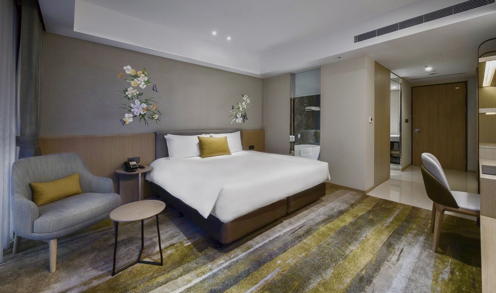 A modern hotel room with a large bed, a seating area with a chair and a small table, and a bathroom visible through an open door.