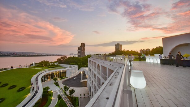 Ein modernes Gebäude mit einer gewölbten Dachform, einem angelegten Garten und Blick auf die Skyline der Stadt bei Sonnenuntergang.