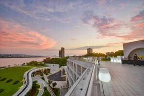 Un bâtiment moderne doté d’un toit incurvé, d’un jardin aménagé et offrant une vue sur les gratte-ciel de la ville au coucher du soleil.
