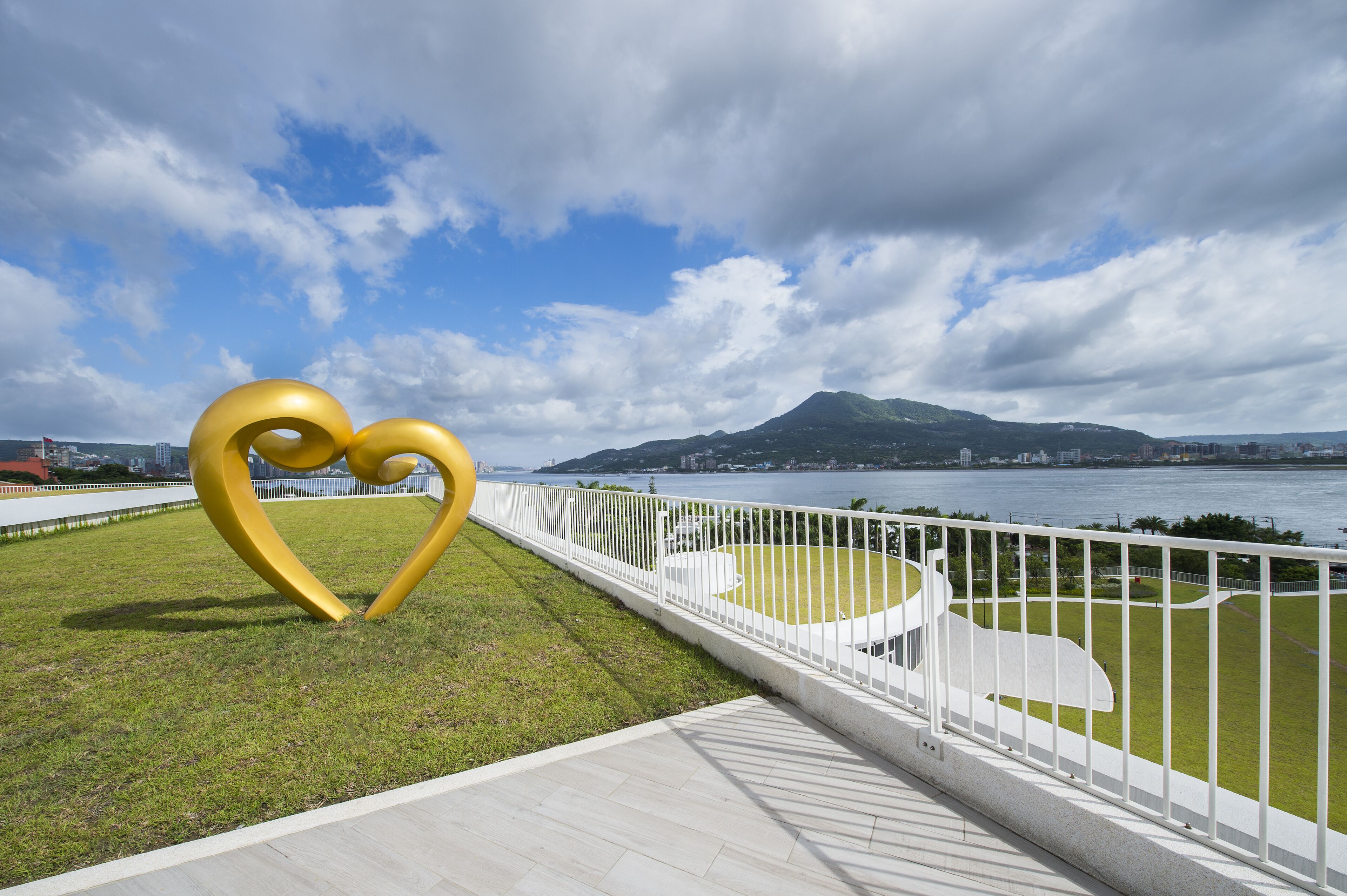 Un parque con una gran escultura dorada en forma de corazón, una valla blanca y una vista de una ciudad y montañas al fondo.