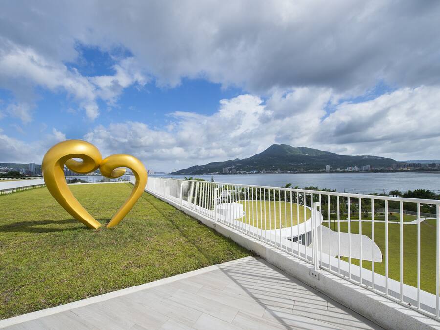 Un parque con una gran escultura dorada en forma de corazón, una valla blanca y una vista de una ciudad y montañas al fondo.