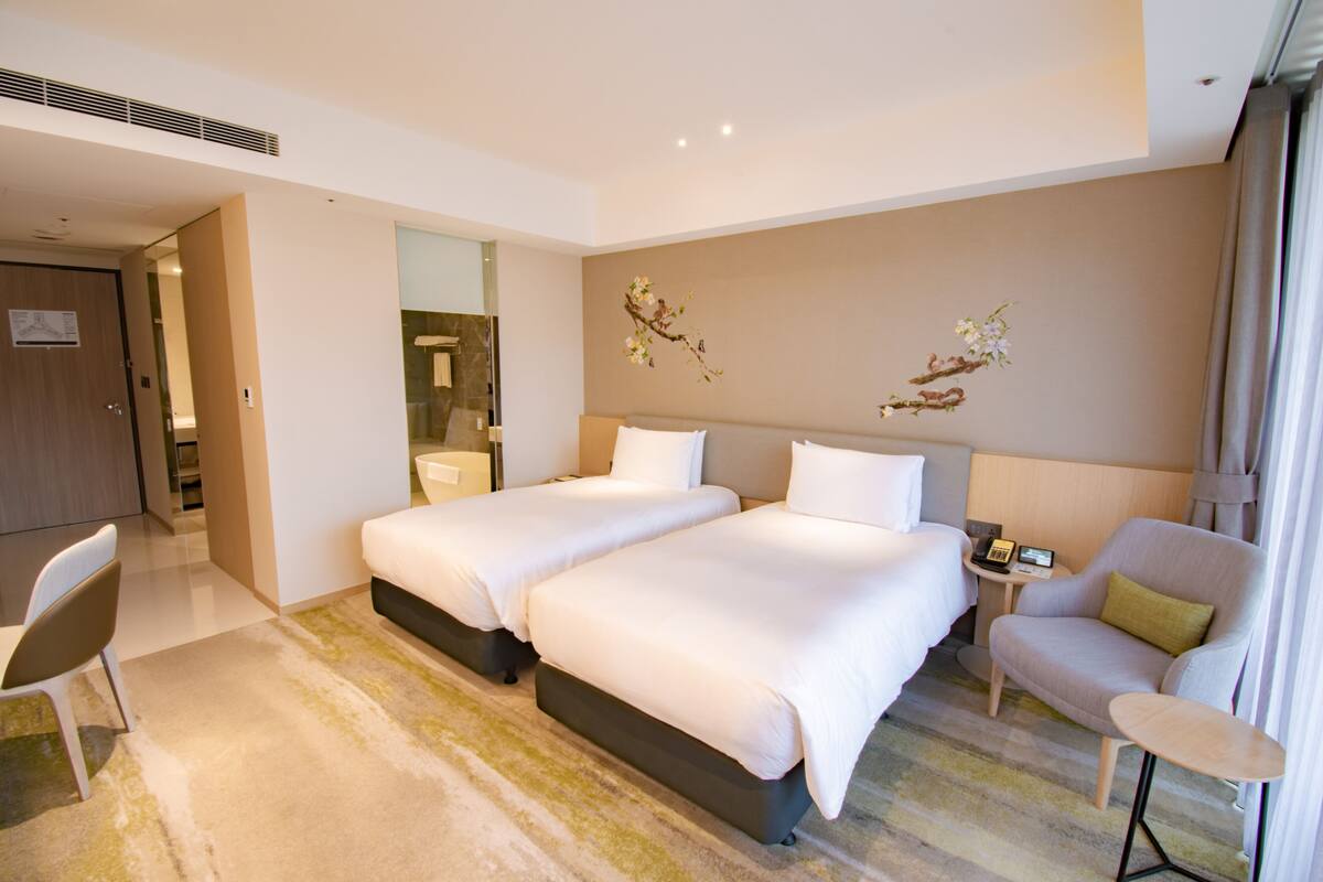 Ein modernes Hotelzimmer mit zwei Betten, einer Sitzecke, einem kleinen Tisch mit Lampe und einem durchgegangene Tür sichtbaren Badezimmer.